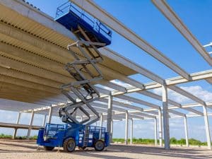 A blue scissor lift, used in ewp training accreditation, is parked on a construction site with steel beams and a concrete roof.