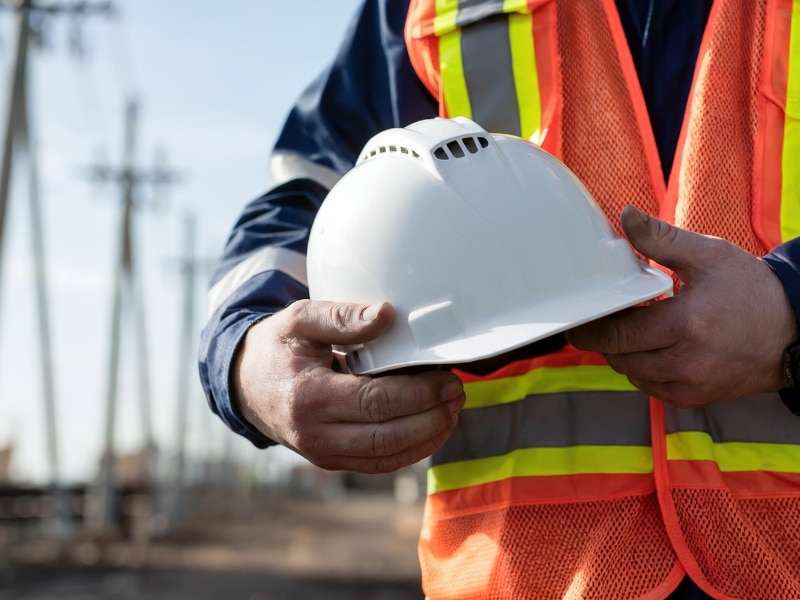 A construction worker in a blue long-sleeved shirt and an orange and yellow high-visibility vest holds a white hard hat in their hands, with a blurred background showing utility poles and a clear sky, prompting the question of **how long to get white card**.
