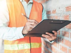 A man wearing a construction vest is writing on a clipboard, with a brick wall in the background. This image relates to the topic of how long to get a white card.